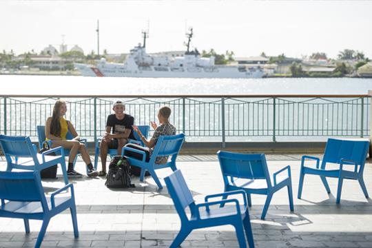 3名学生在阿罗哈塔码头附近的蓝色椅子上聊天. 在他们身后，一条船在水里.